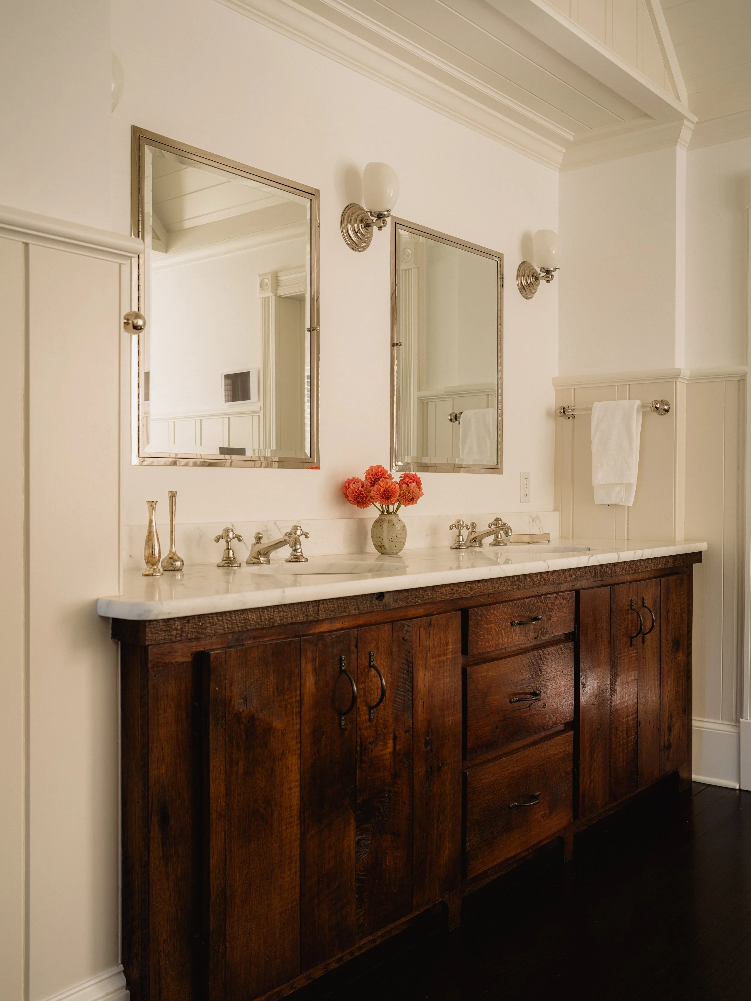 Bathroom vanity with a dark wooden cabinet, a white marble countertop, two mirrors, and two wall-mounted lights. A vase with red flowers is on the countertop, and there is a towel on a towel bar to the right.