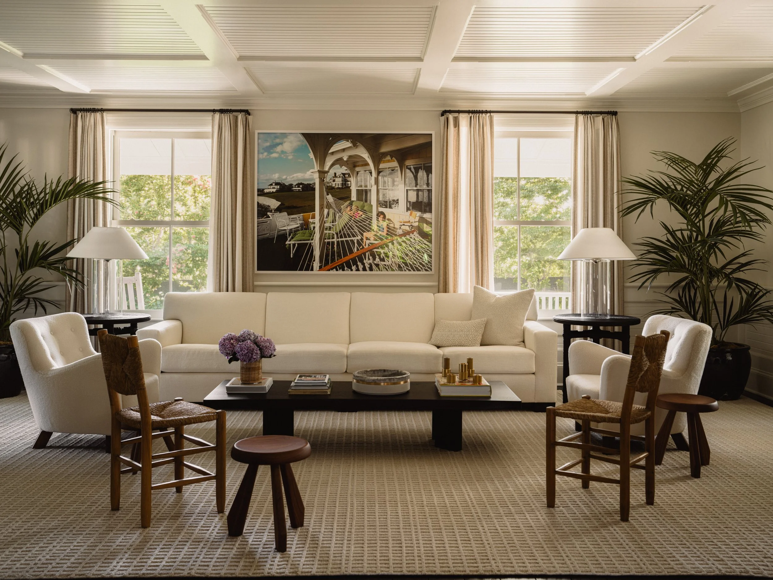 Living room with beige sofa, two armchairs, several wooden chairs, coffee table with flowers, large windows with curtains, house photo on wall, potted plants, and lamps.