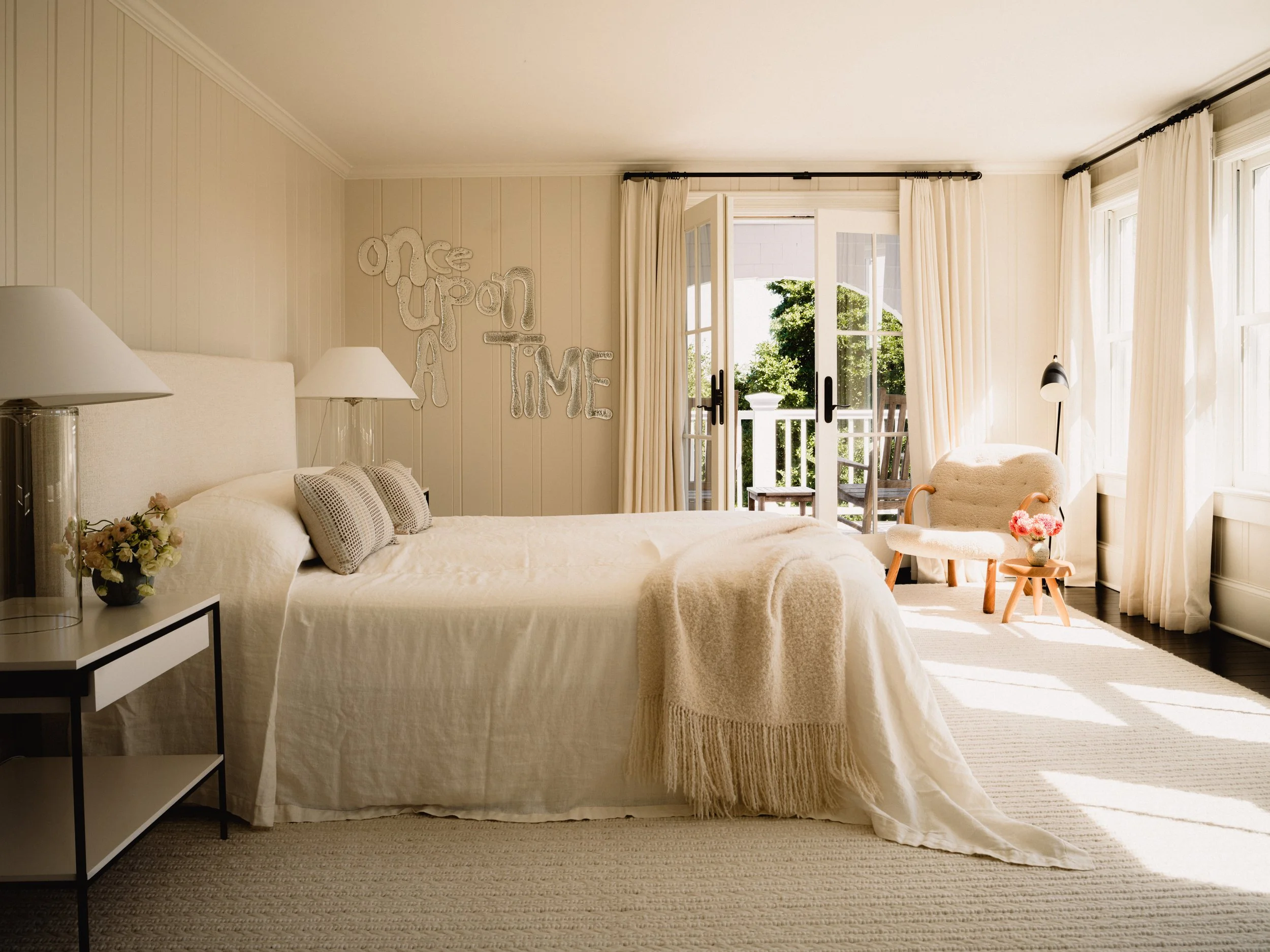 A bedroom with a cream-colored bed, bedside lamps, a vase of flowers, a small sculpture, large windows with cream curtains, a balcony with green trees outside, and a wall decoration that reads 'one upon a time'.