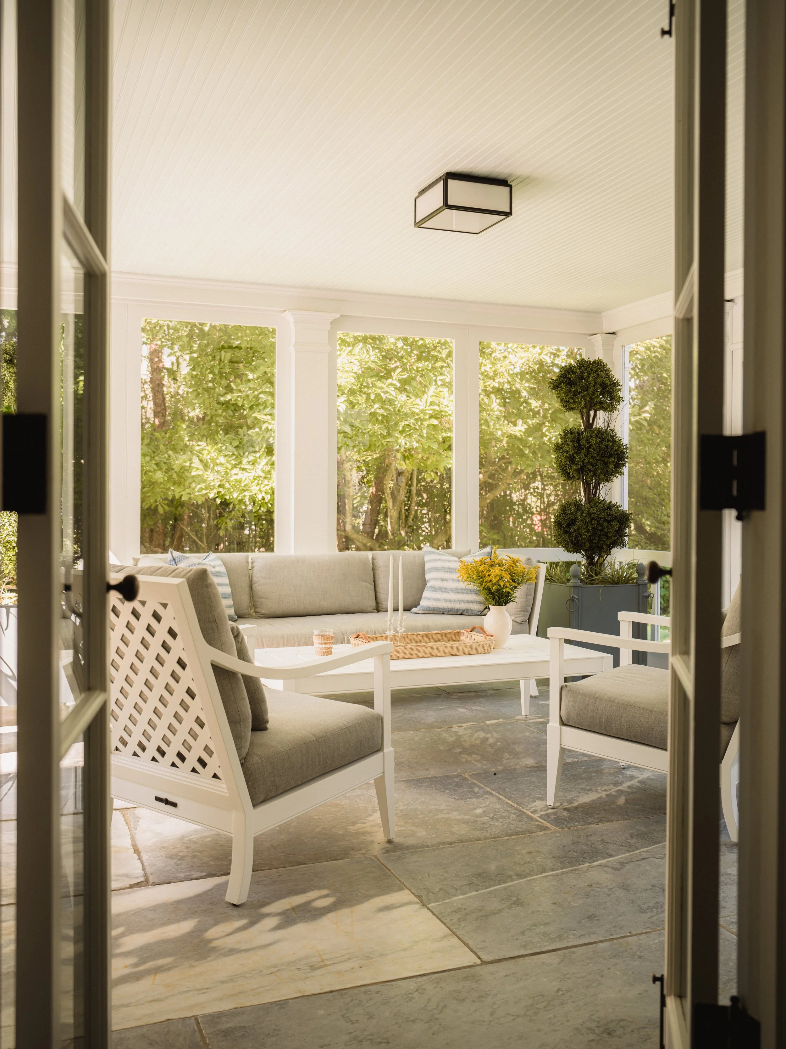 View through open door into a sunny porch with white furniture, a sofa, white chairs, plants, and a table, overlooking a green garden.