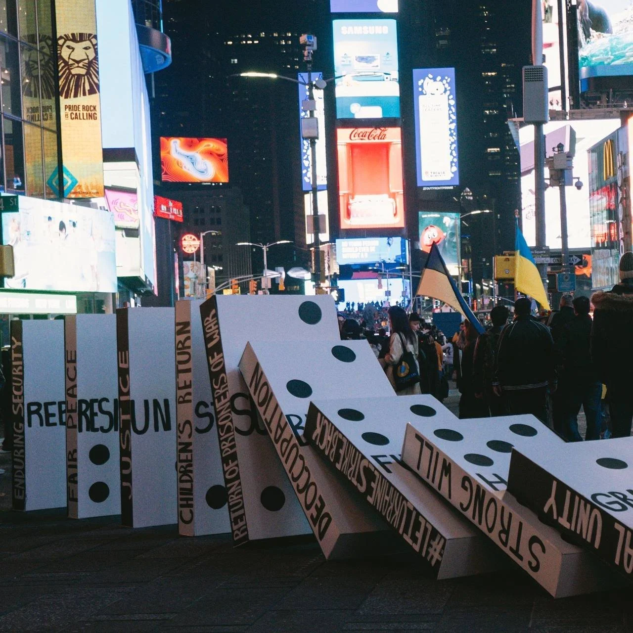 Times Square at night with bright billboards and screens. People holding Ukrainian flags. A large broken domino sculpture with words like 'Resilience', 'Children', and 'Peace'
