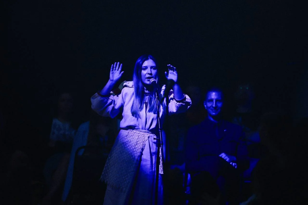 Woman speaking or singing into a microphone on stage, with hands raised, illuminated in blue light, audience members visible in the background.