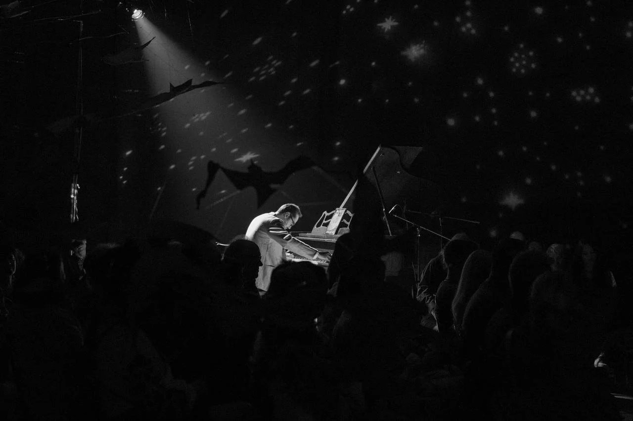 A pianist performing on stage with star and bat shaped shadows projected on the ceiling, an audience watching in the dark.