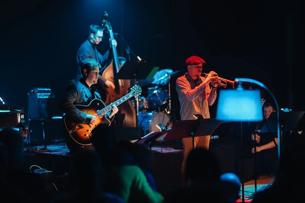 Musicians performing on stage, with a guitarist, a trumpet player, and a double bassist, under dim lighting.