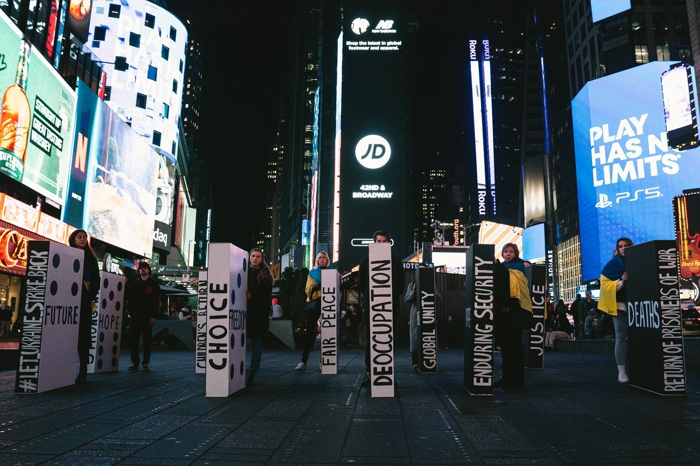 Protesters holding signs with messages such as 'Future', 'Hope', 'Choice', 'Fair', 'Peace', 'Decaffeination', 'Global Unity', 'Enduring Security', 'Justice', 'Deaths', and 'Return of Prisoners', in Times Square at night with bright digital billboards
