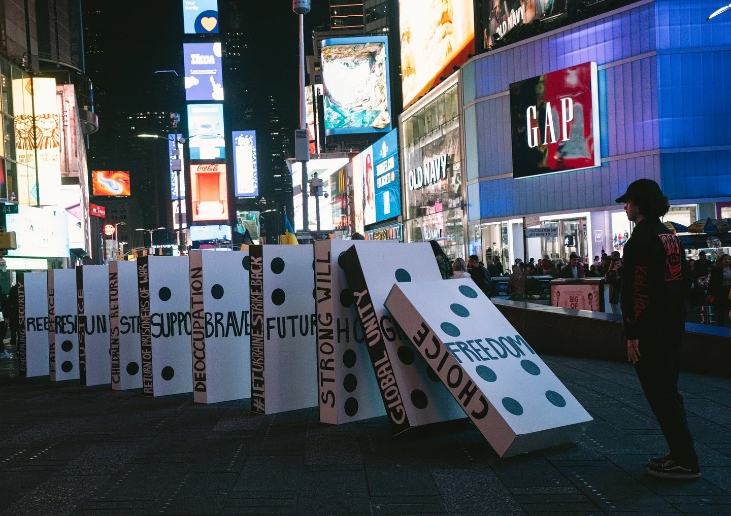 Nighttime scene in Times Square, New York City, with illuminated digital billboards and advertisements. In the foreground, a display mimics dominoes with words like 'RESIST,' 'SUPPORT,' 'FUTURE,' and 'FREEDOM,' some dominoes knocked over. A man in da