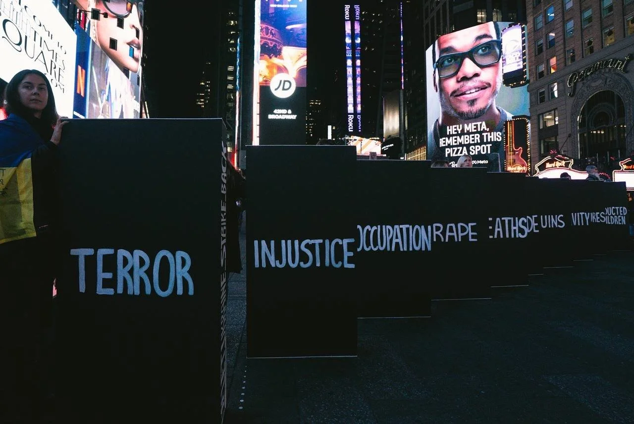 Protest setup with black panels displaying words like 'TERROR', 'INJUSTICE', 'OCCUPATION', 'RAPE', 'VITRIOLS', and 'DESTRUCTION'. Large digital billboards in the background showing advertisements and a person with glasses on one screen. A woman holdi