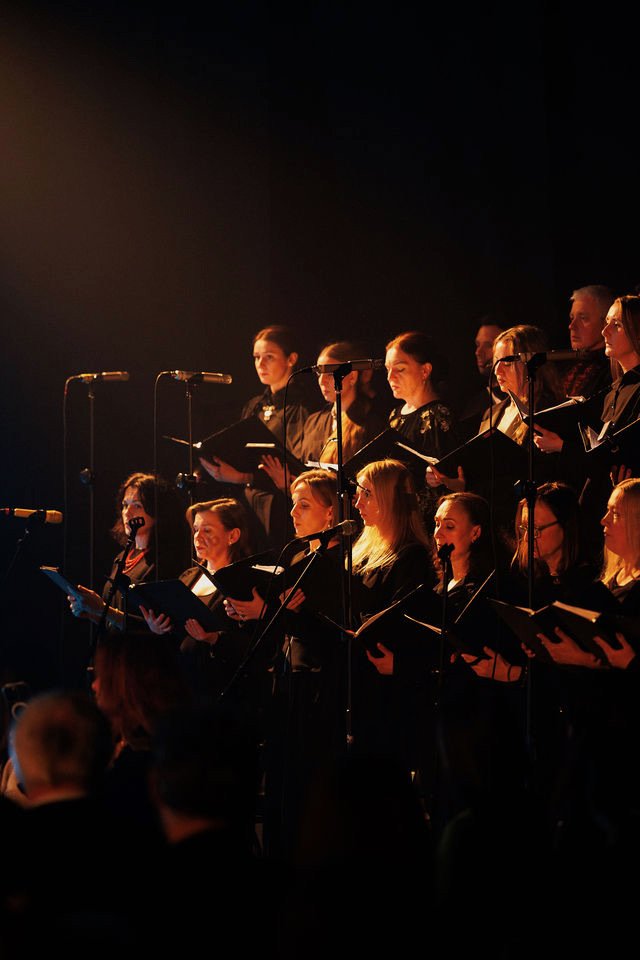 A choir of women dressed in black performing on stage, singing from sheet music, with microphones set up in front of them.