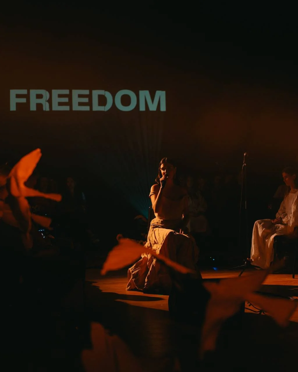 A woman sits on a stage with a microphone, illuminated by warm lighting, with the word 'FREEDOM' projected behind her. There are other people sitting on the stage and the audience is visible in the dark.