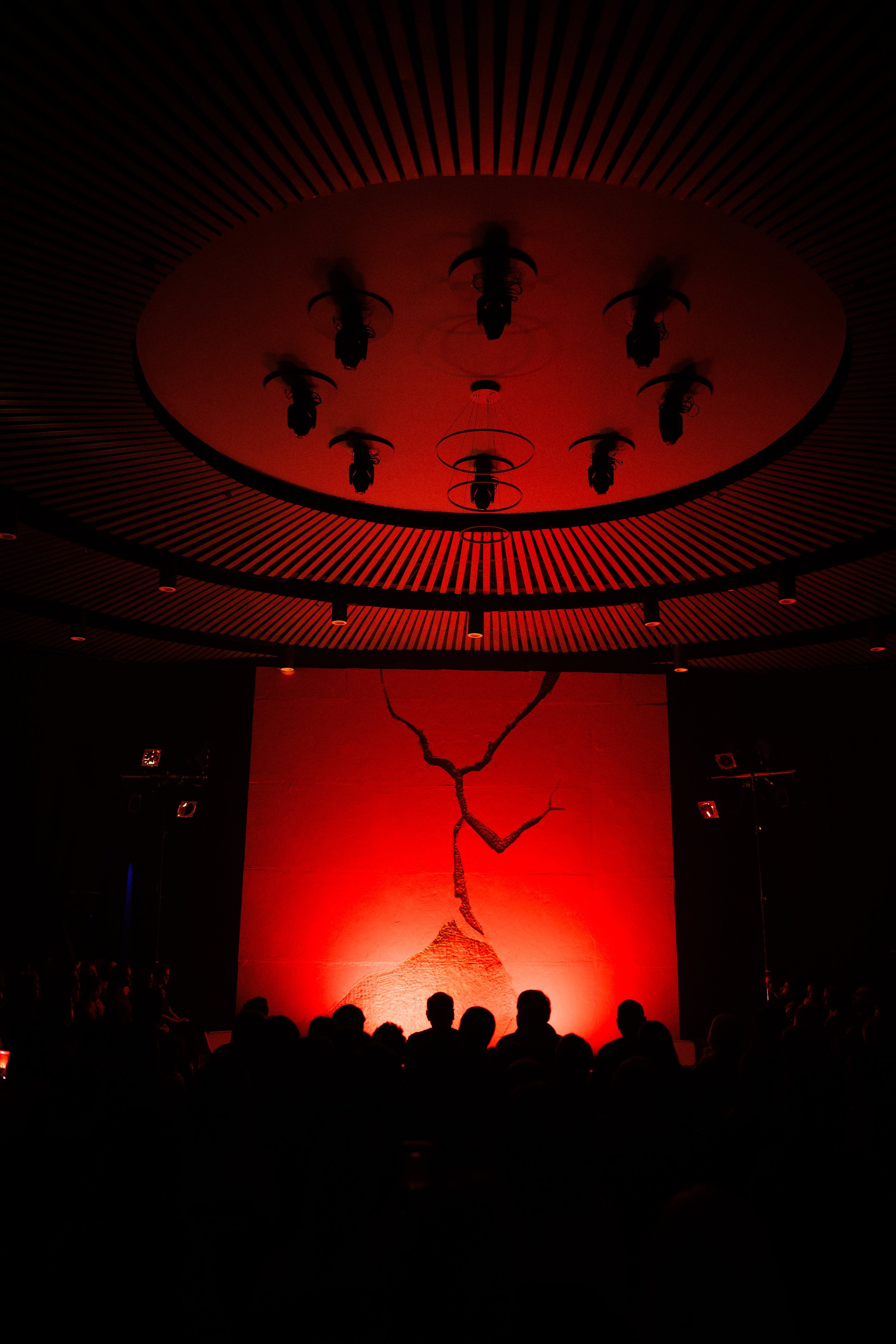 Darkened auditorium with a large red-lit wall featuring a cracked mountain and sunset image, silhouettes of audience members and ceiling lights with circular fixtures.