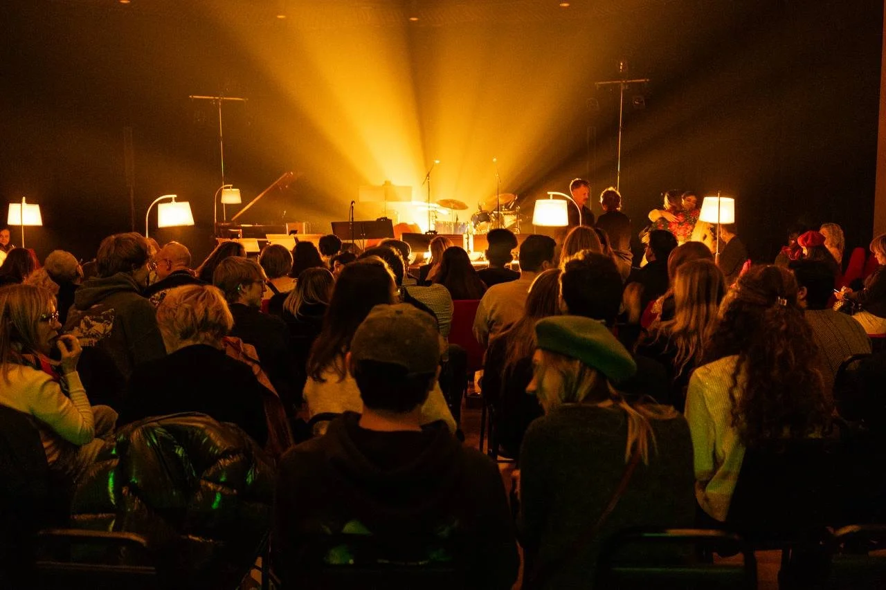 Audience seated in a dimly lit concert venue, awaiting a performance. The stage has a few illuminated lamps and musical instruments, with warm yellow lighting radiating from behind.