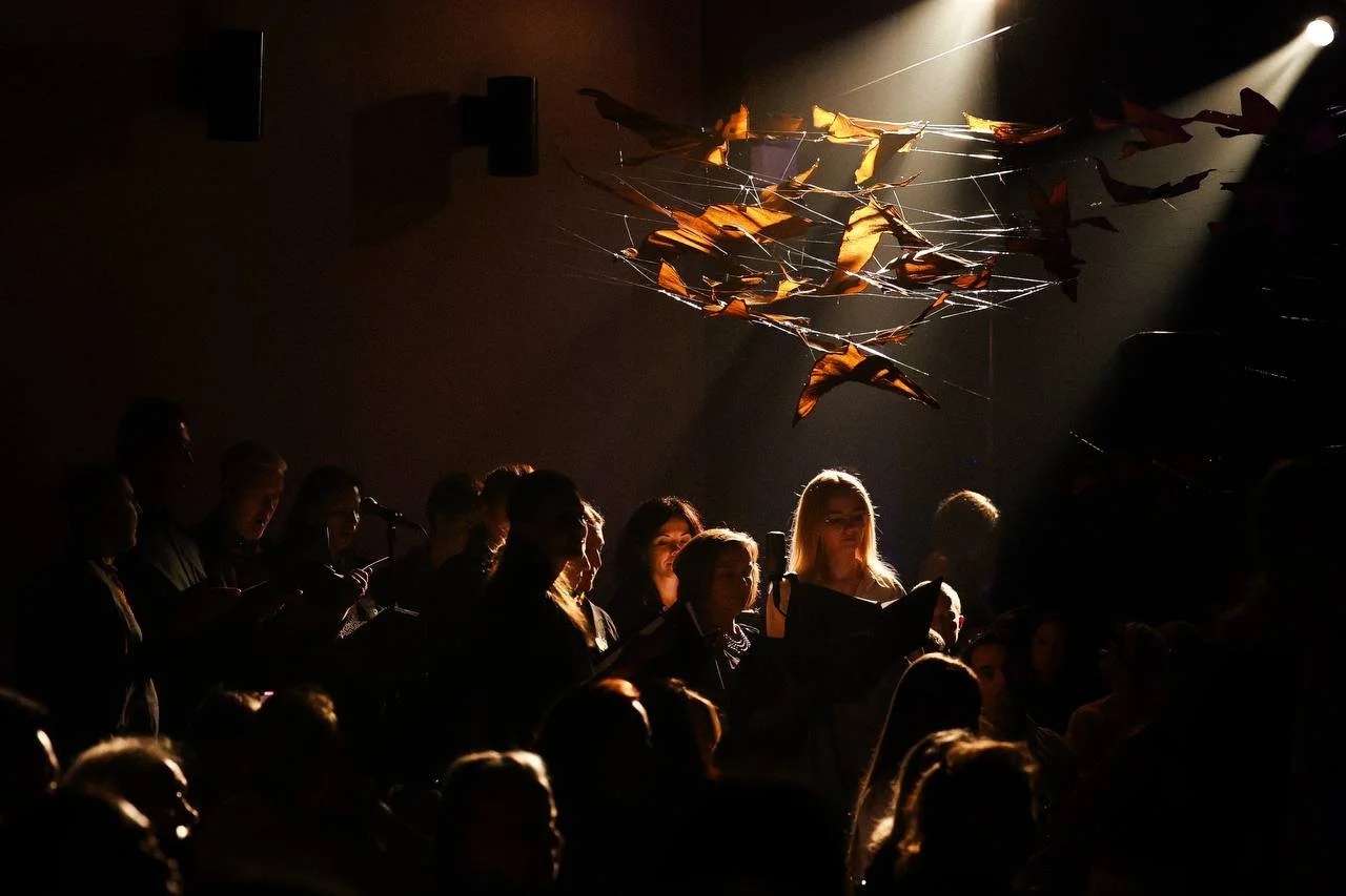 A choir singing in a dimly lit concert hall with a dramatic spotlight shining on them, and an art installation hanging above with orange fabric pieces and web-like metal structure.