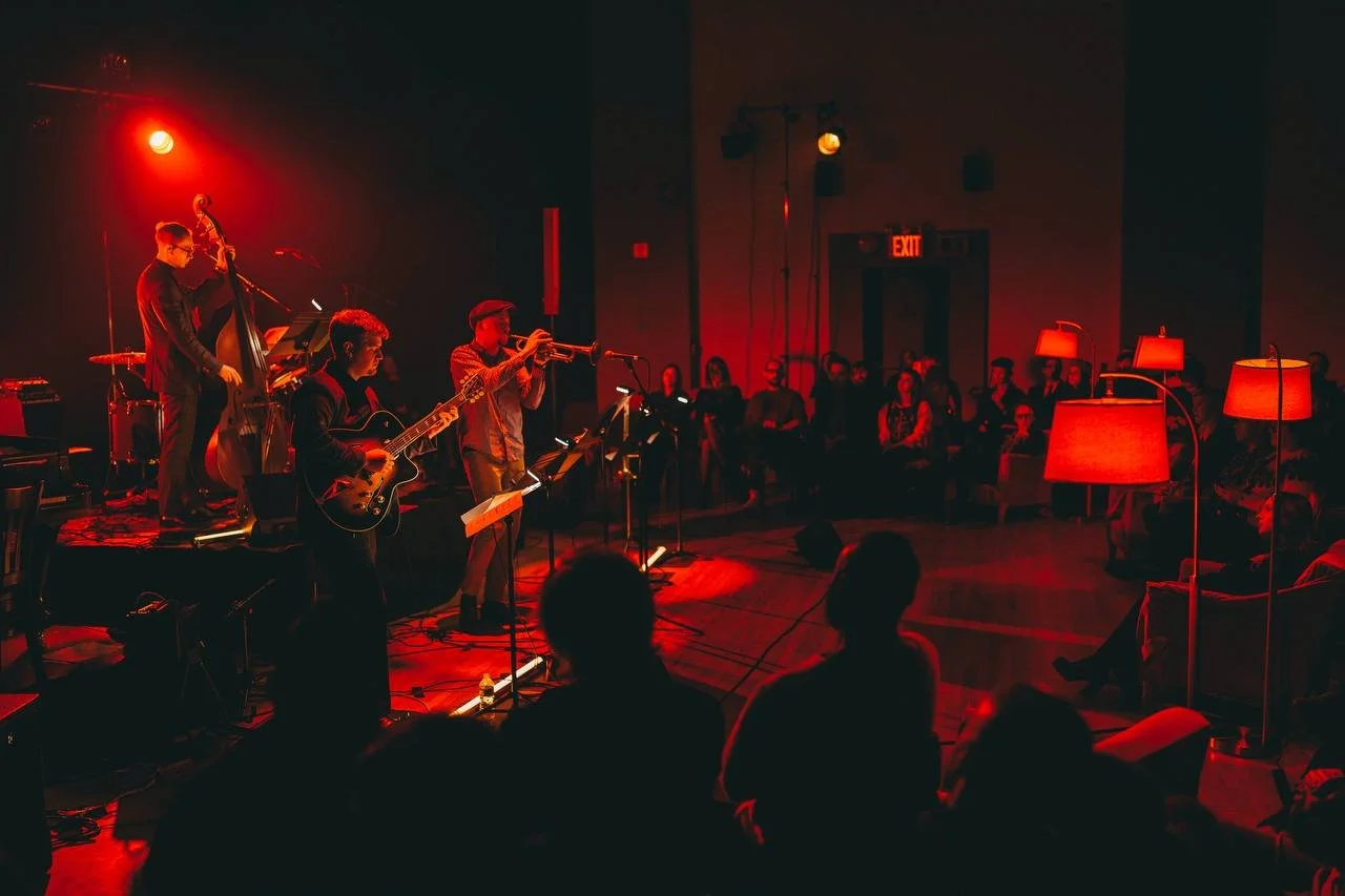 A jazz band performs on stage with red lighting, featuring musicians playing guitar, trumpet, and upright bass, with an audience seated and watching in a dimly lit room.