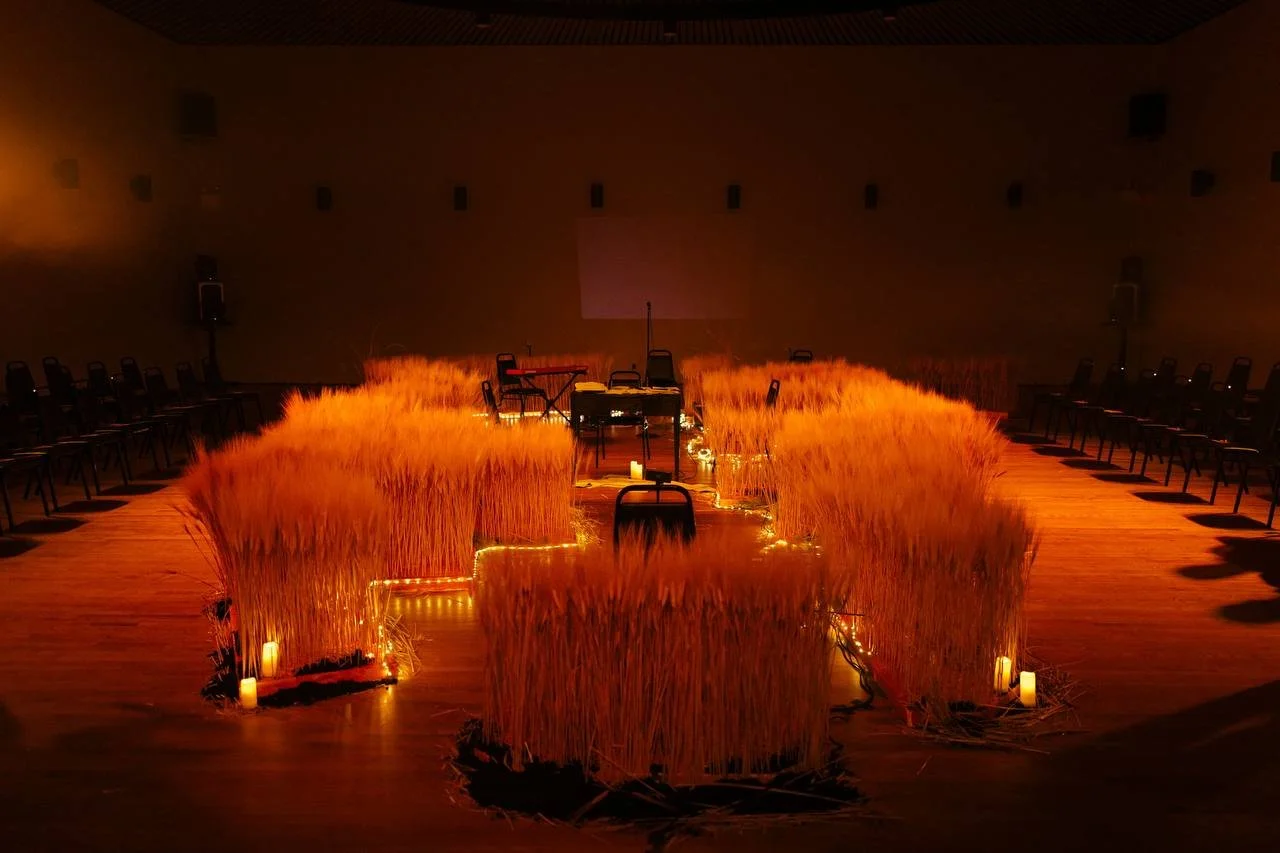 Empty conference room decorated with tall, golden wheat stalks and candles arranged in a U-shape, with chairs placed around the perimeter and musical instruments at the front.