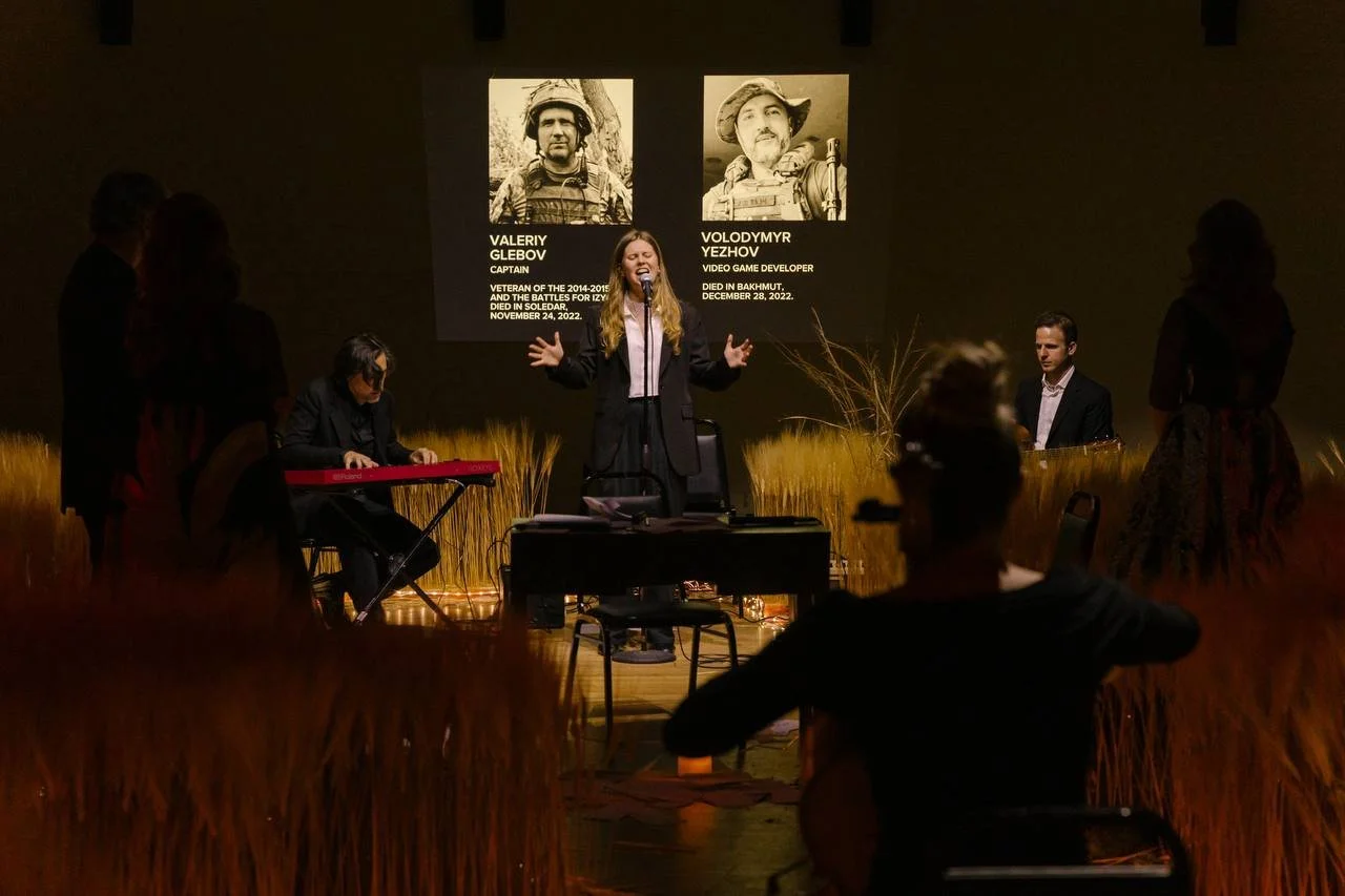 A woman is standing on stage, speaking into a microphone during a memorial event. Behind her are large portraits and biographical descriptions of Valeriy Glebov and Volodymyr Yezyhov. There are people seated and standing around the stage, with a keyb