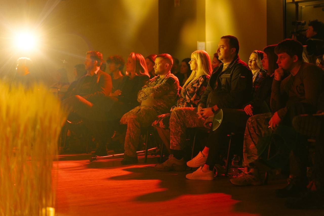 Audience seated in darkened room with warm lighting, watching a performance.