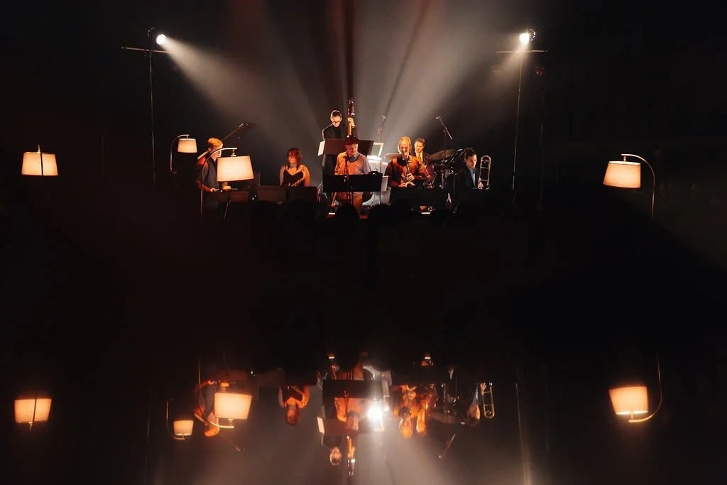 A jazz band performing on a dark stage with lamps and a reflective surface below, creating a mirror image of the band members.