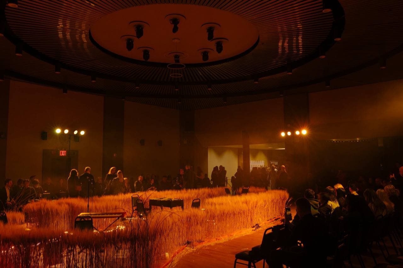 An indoor event space with a stage surrounded by tall, golden grass-like plants, with people gathered around. There are multiple bright spotlights directed towards the stage area, and a large ceiling fixture. Some people are sitting, while others are