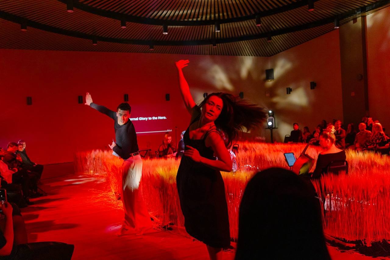 Two dancers perform on a stage with a red illuminated backdrop, surrounded by wheat-like plants. Audience members sit on either side, some capturing the performance on their phones. The scene is vibrant with dynamic lighting.