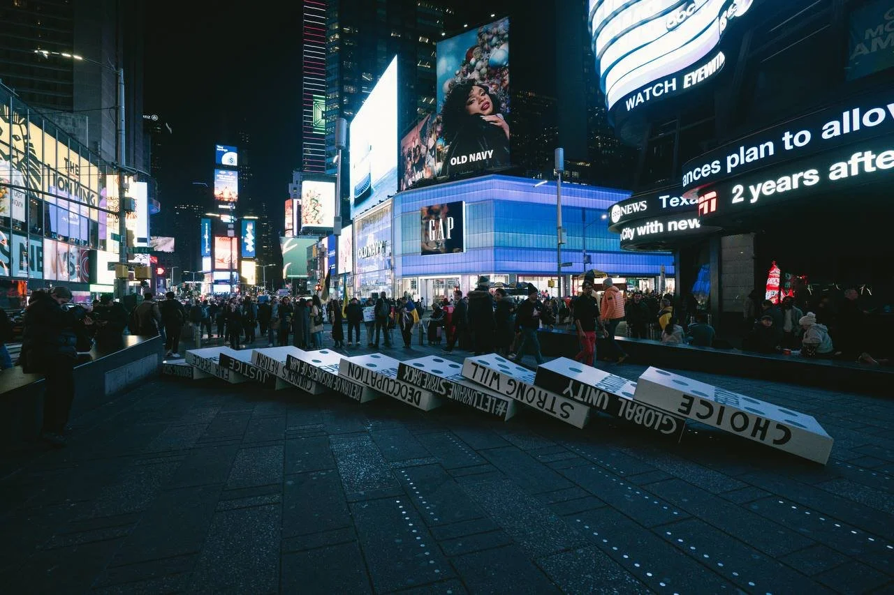 Nighttime scene in Times Square with digital billboards, LED screens, and crowds of people, some standing and walking around. There is a toppled sign on the ground that reads "CHOICE" and other words. The area is illuminated by bright lights from adv