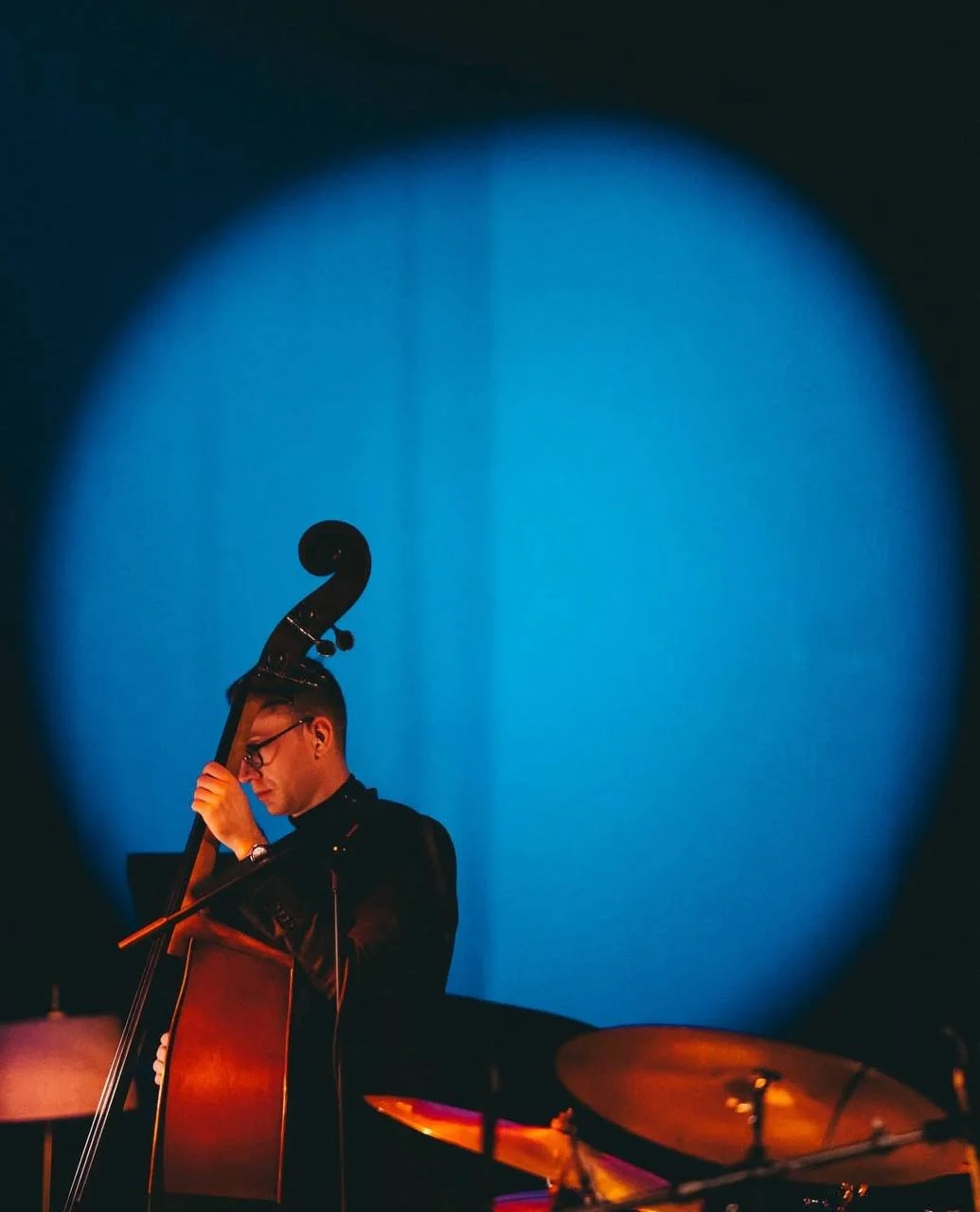 A man wearing glasses playing a double bass on a dimly lit stage with a blue spotlight behind him.