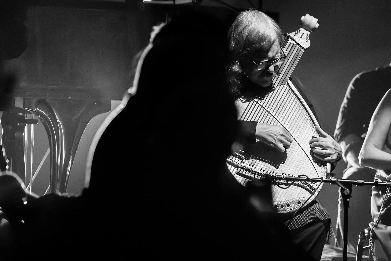 A musician playing a hammered dulcimer on stage, with other performers partially visible, in black and white.