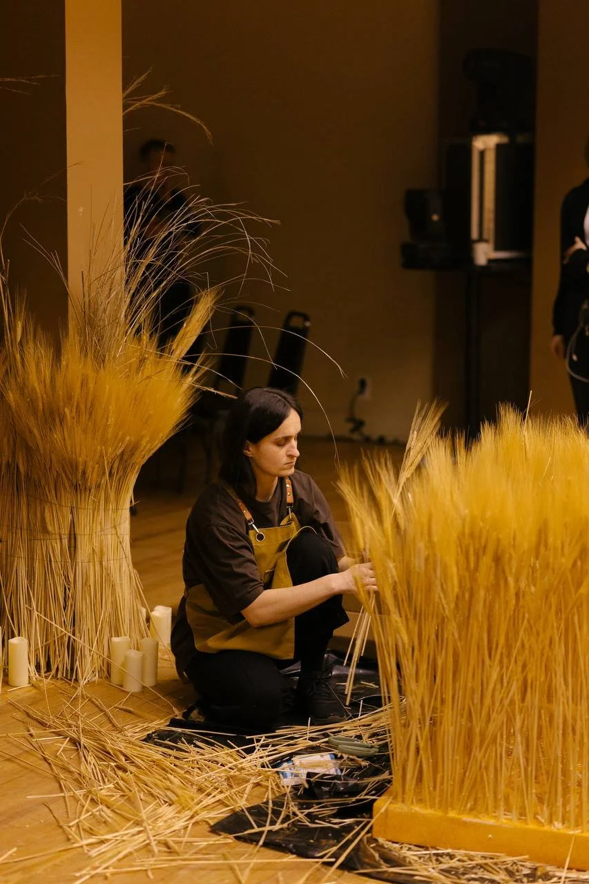 A woman sitting on the floor surrounded by wheat stalks, arranging tall wheat stalks in a display or craft project in an indoor setting.