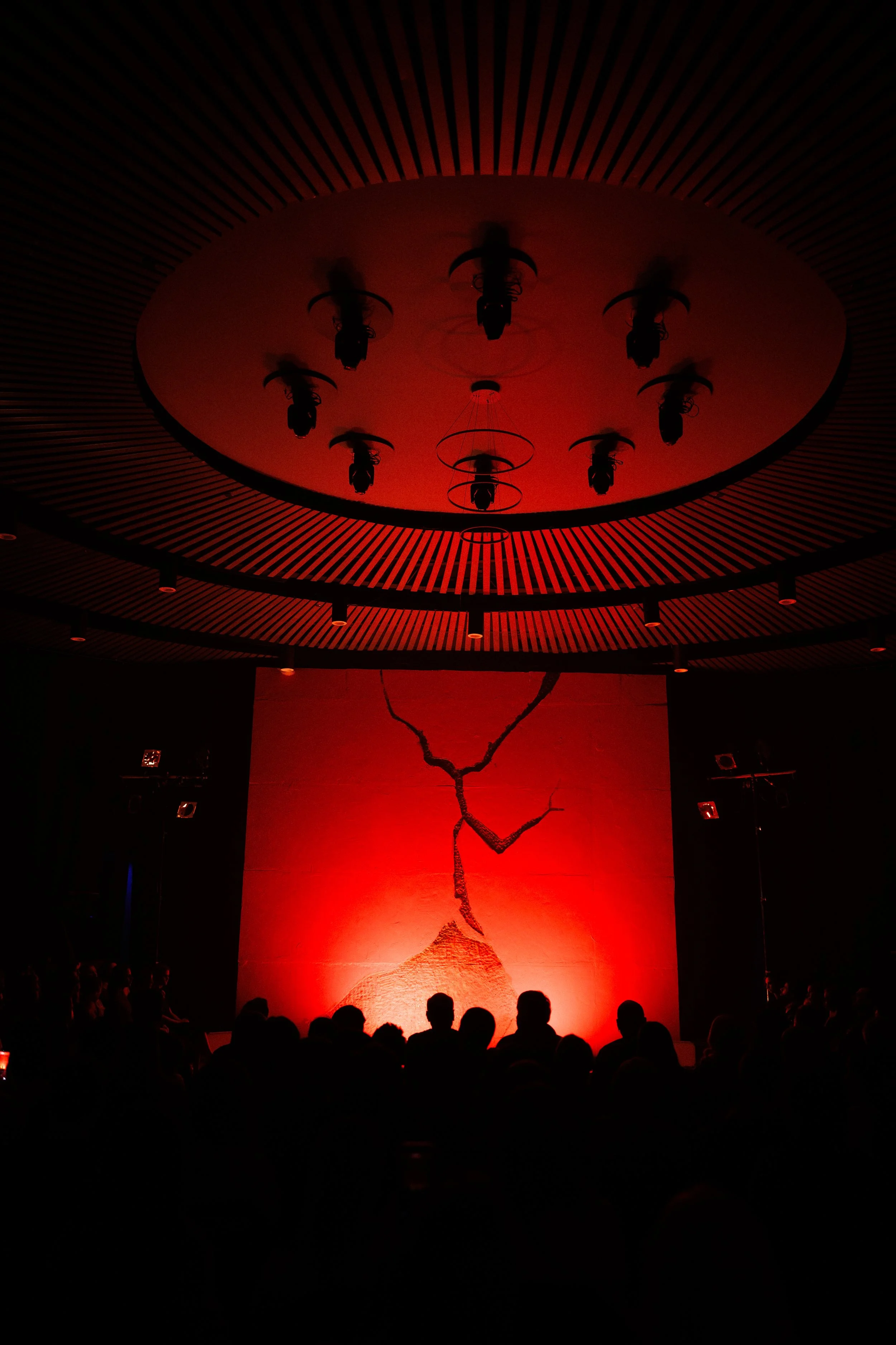 A dark auditorium with a stage illuminated in red, featuring a cracked mountain of some sort painting or sculpture with silhouettes of audience members in the foreground.