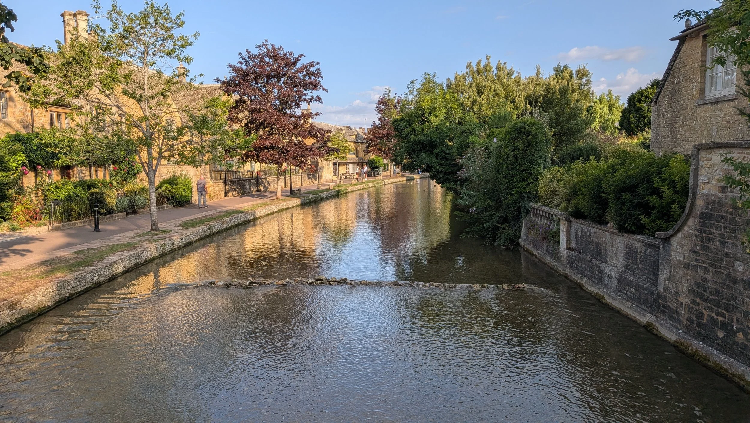 A scenic view of a small river or canal running through a picturesque town with stone buildings, lush green and red-leafed trees, and a walking path along the water