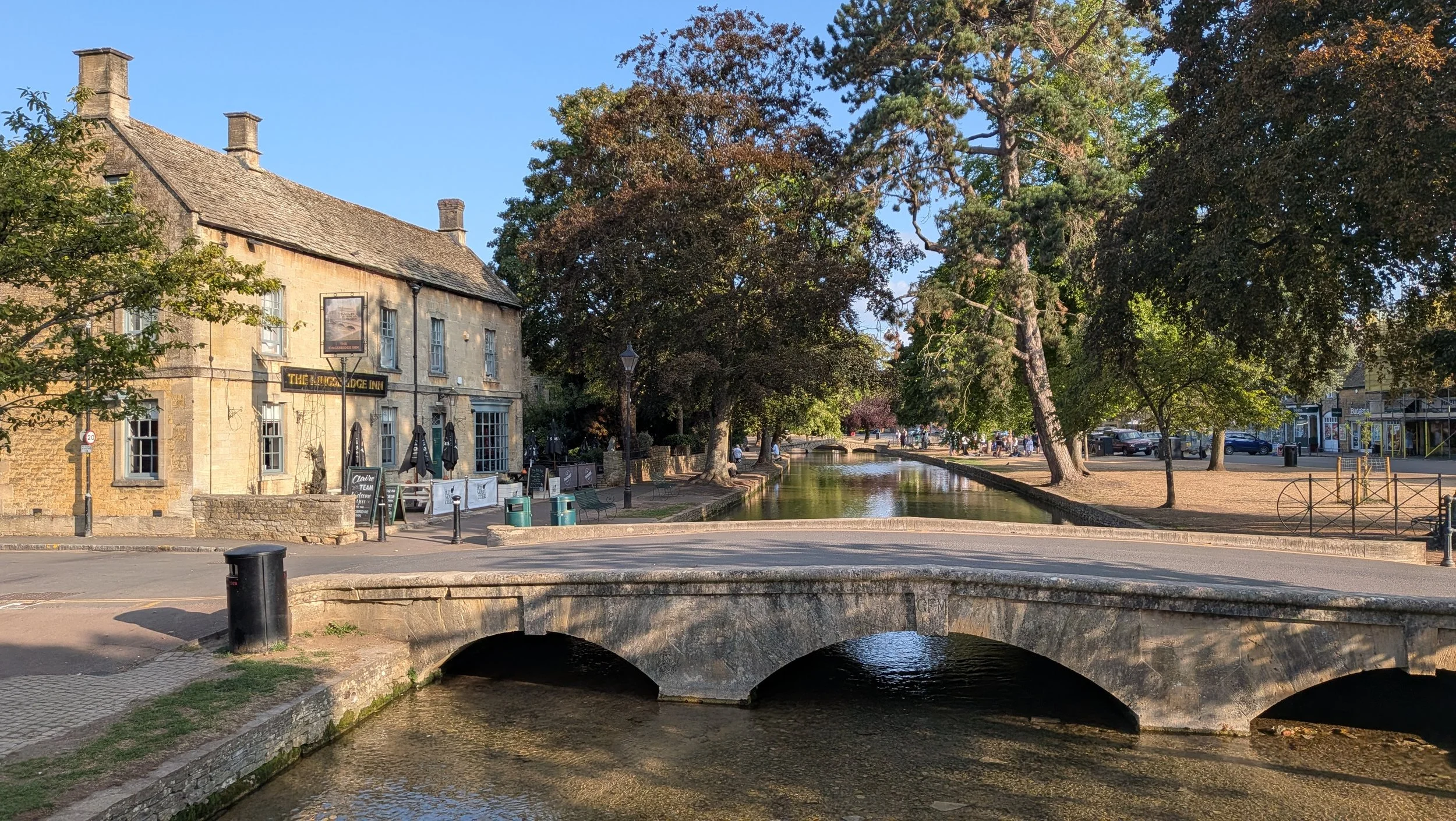 A canal with a small stone bridge in the foreground, flanked by a building on the left and a park with trees on the right. Outdoor seating and a few people are visible in the background, under a clear blue sky.