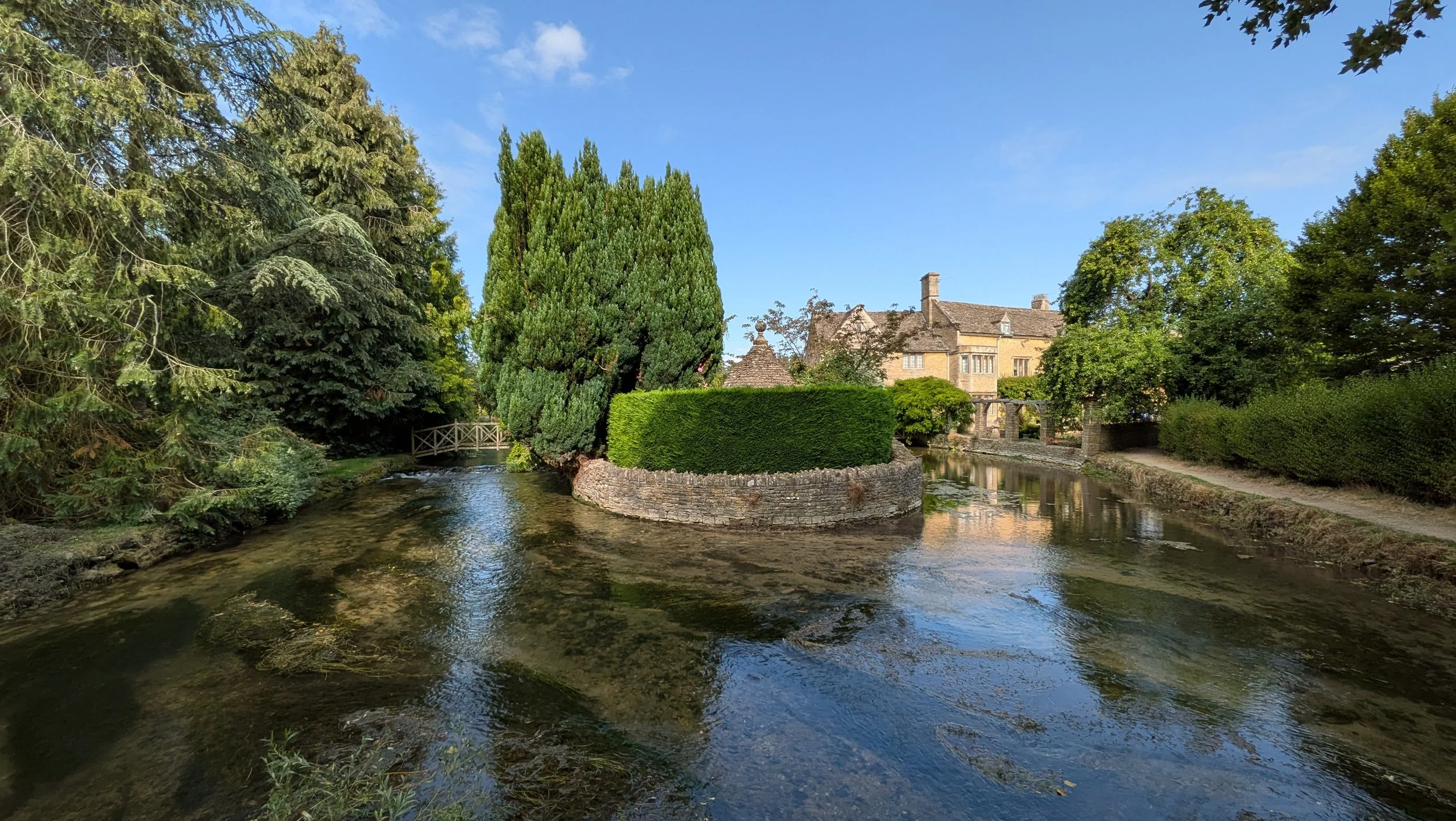 A scenic view of a small canal with clear water, surrounded by lush green trees and shrubs. There is a stone building with a sloped roof in the background, and a small bridge crossing over the canal on the left side of the image.
