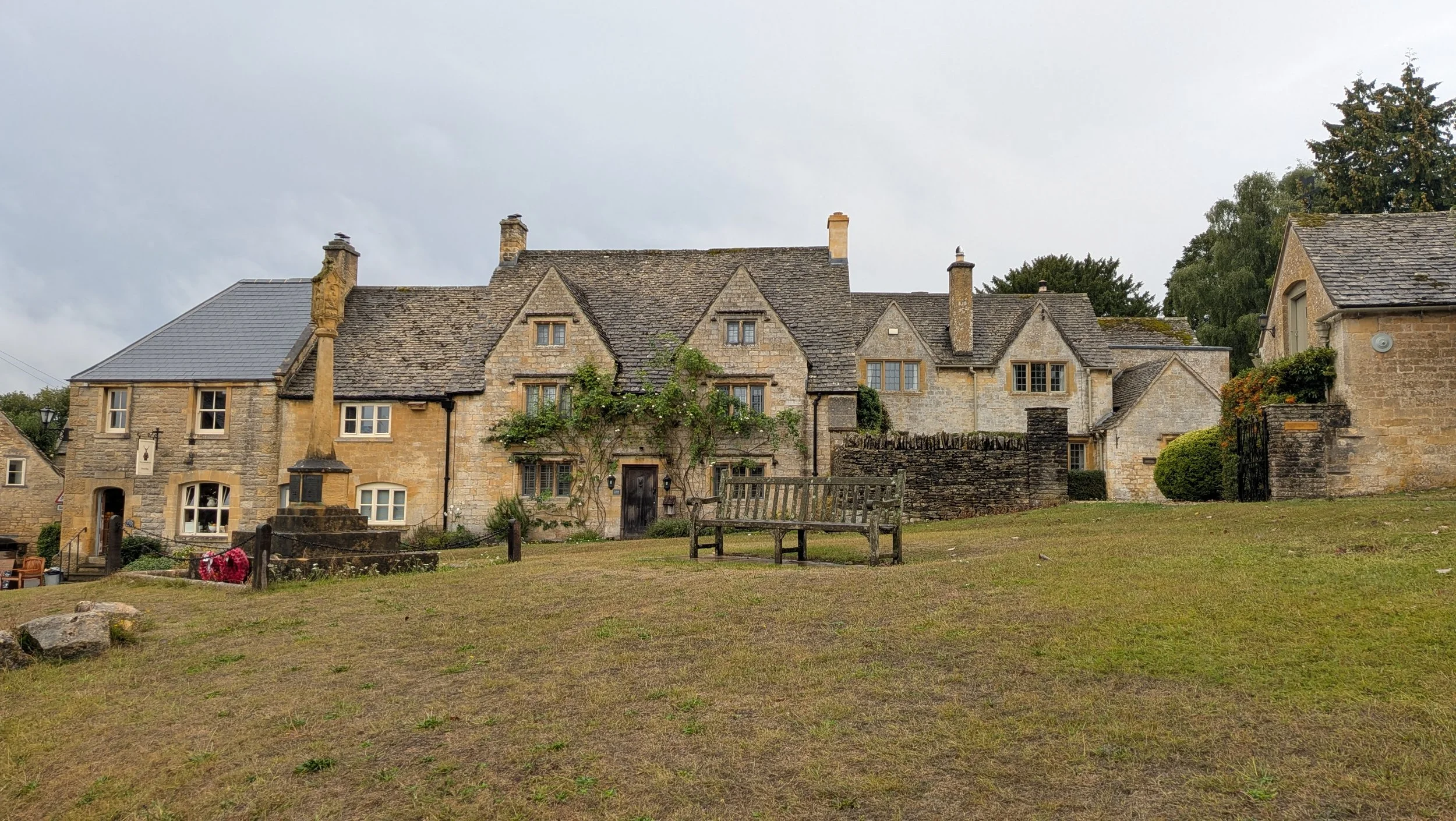 A historic stone building with multiple gabled roofs, chimneys, and windows, surrounded by a lawn and some trees under a cloudy sky.