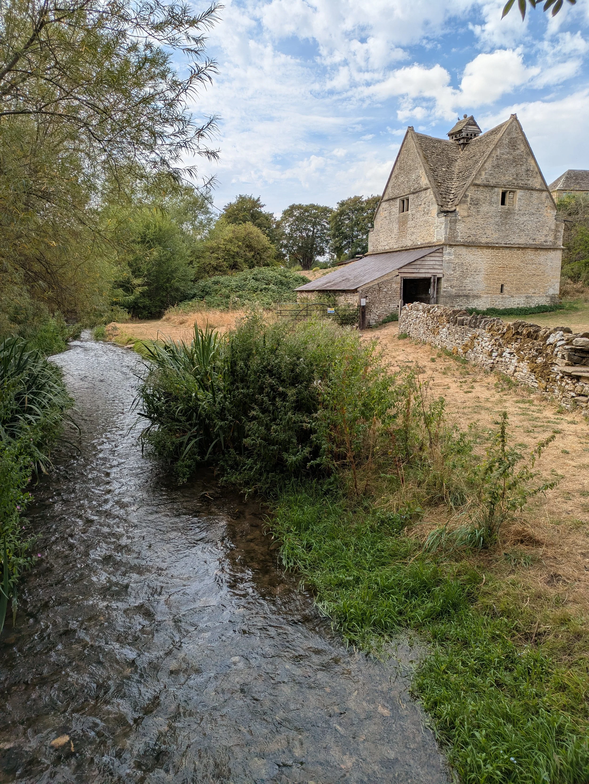 A historic stone building with a steep roof and small windows, situated next to a small stream with green foliage and trees around, under a partly cloudy sky.
