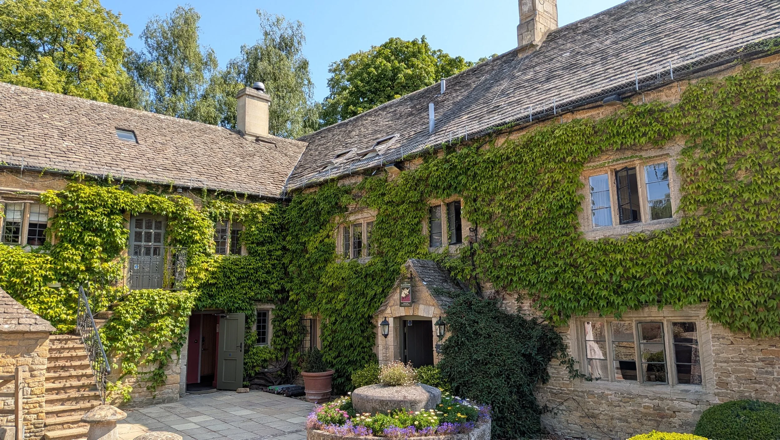 A stone house covered in green ivy with several windows, a door, and a small garden with flowers in a pot, under a clear blue sky.