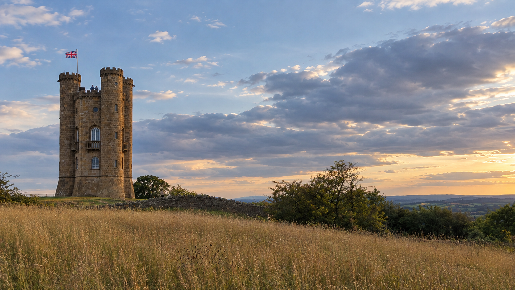 A historic stone castle tower topped with a Union Jack flag, standing on a grassy hill against a cloudy sunset sky, with rolling green hills and trees in the background.