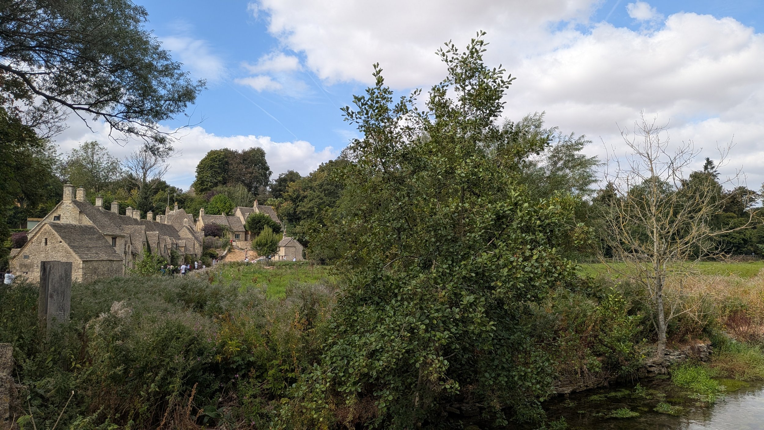 View of an old village with stone cottages and chimneys, surrounded by lush greenery and trees, under a partly cloudy sky.