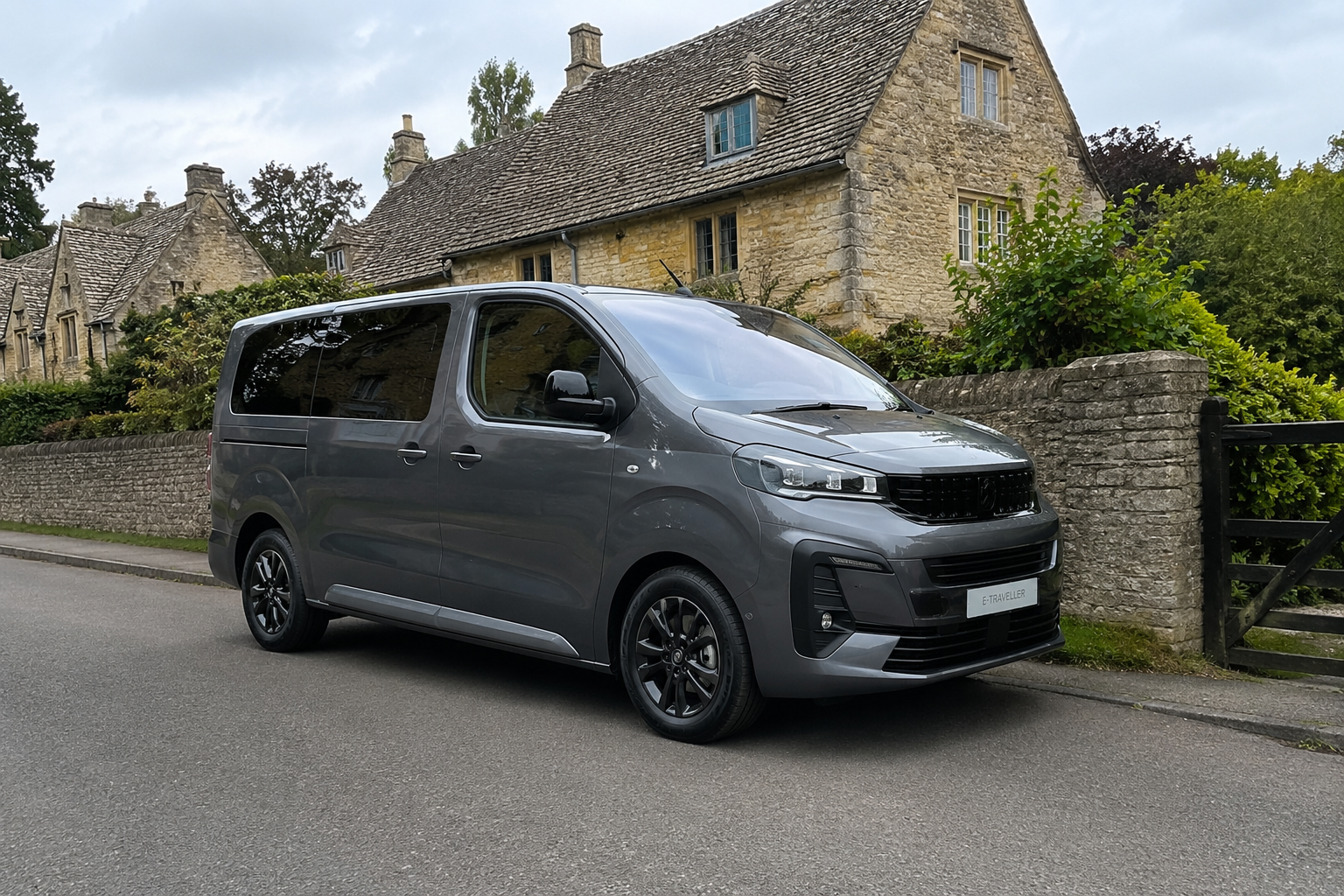 A modern gray electric van parked on a residential street in front of stone houses and a stone wall with greenery.