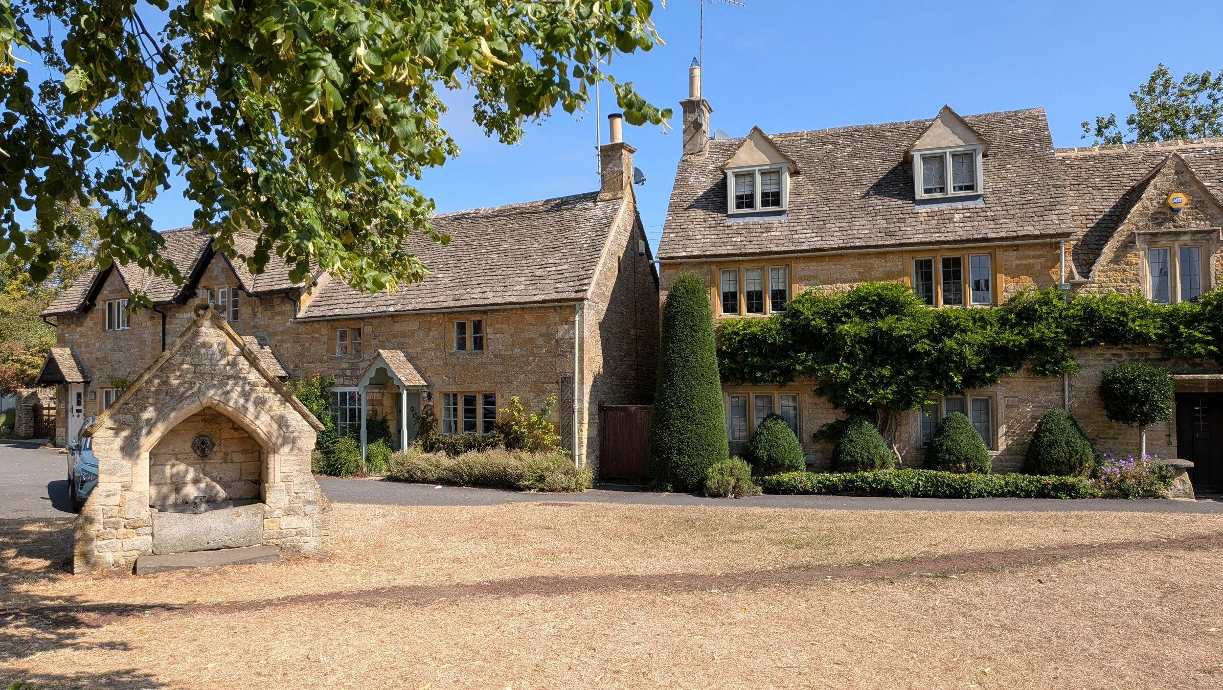 A stone house with dormer windows and a pitched roof, surrounded by manicured bushes and trees under a blue sky.
