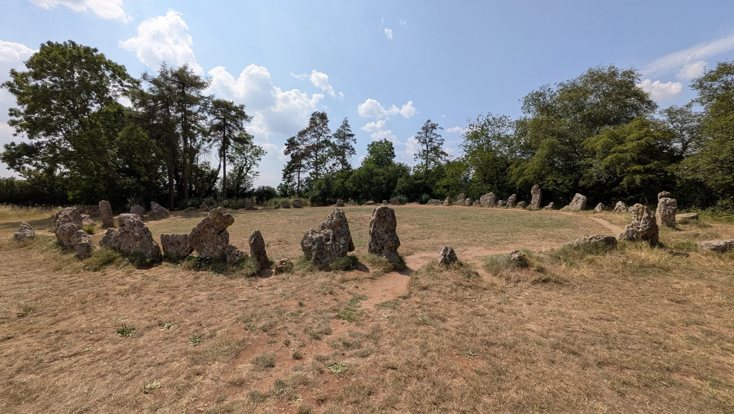 A circular arrangement of upright stones in a grassy area with trees and a partly cloudy sky in the background.