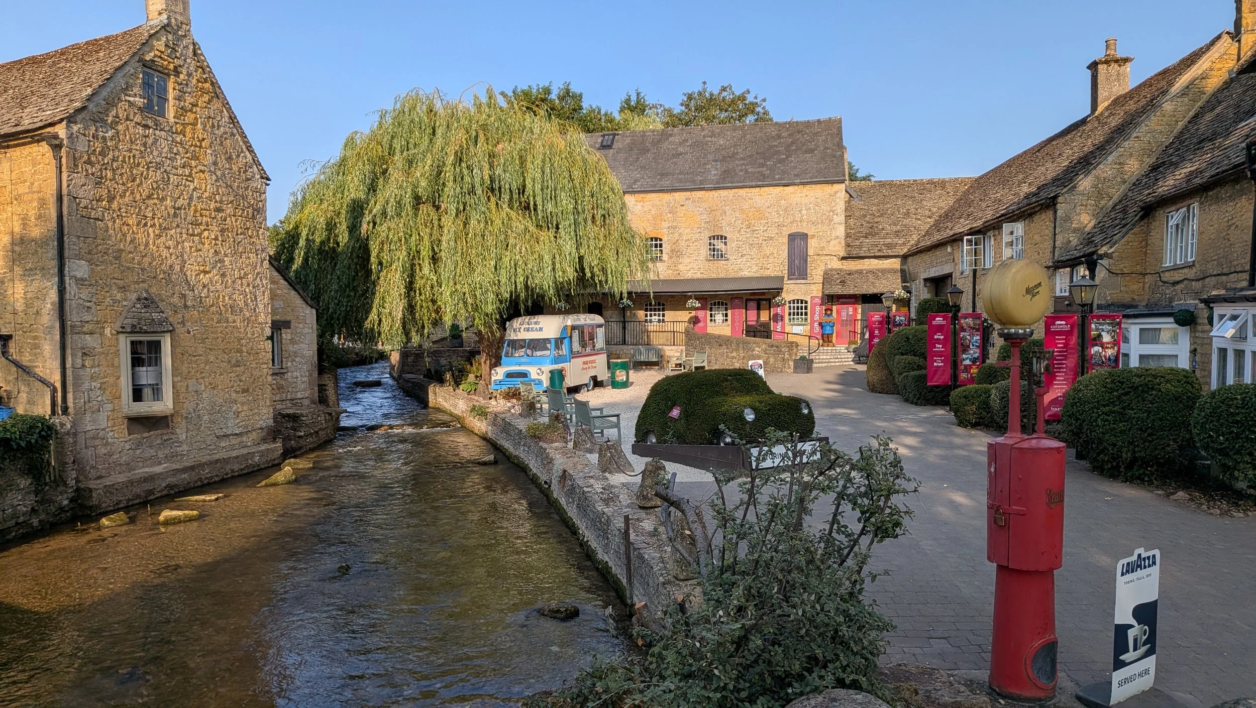 A scenic small town street with old stone buildings, a small stream, a large leafy tree, a vintage ice cream truck, benches, red informational signs, and a red postbox under a clear blue sky.