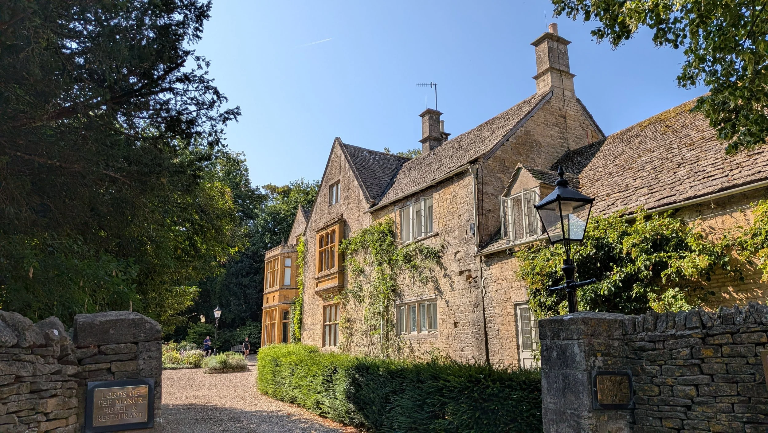 A stone historic building with large windows and a steep roof in a garden setting, surrounded by trees and a stone wall with a sign reading 'Lords of The Manor Hotel & Restaurant'.