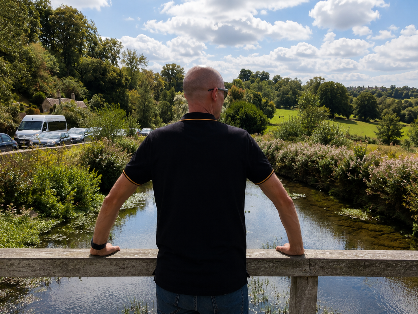 A man wearing sunglasses and a black polo shirt standing on a wooden bridge, overlooking a small river with lush green trees and hills in the background on a bright, partly cloudy day.