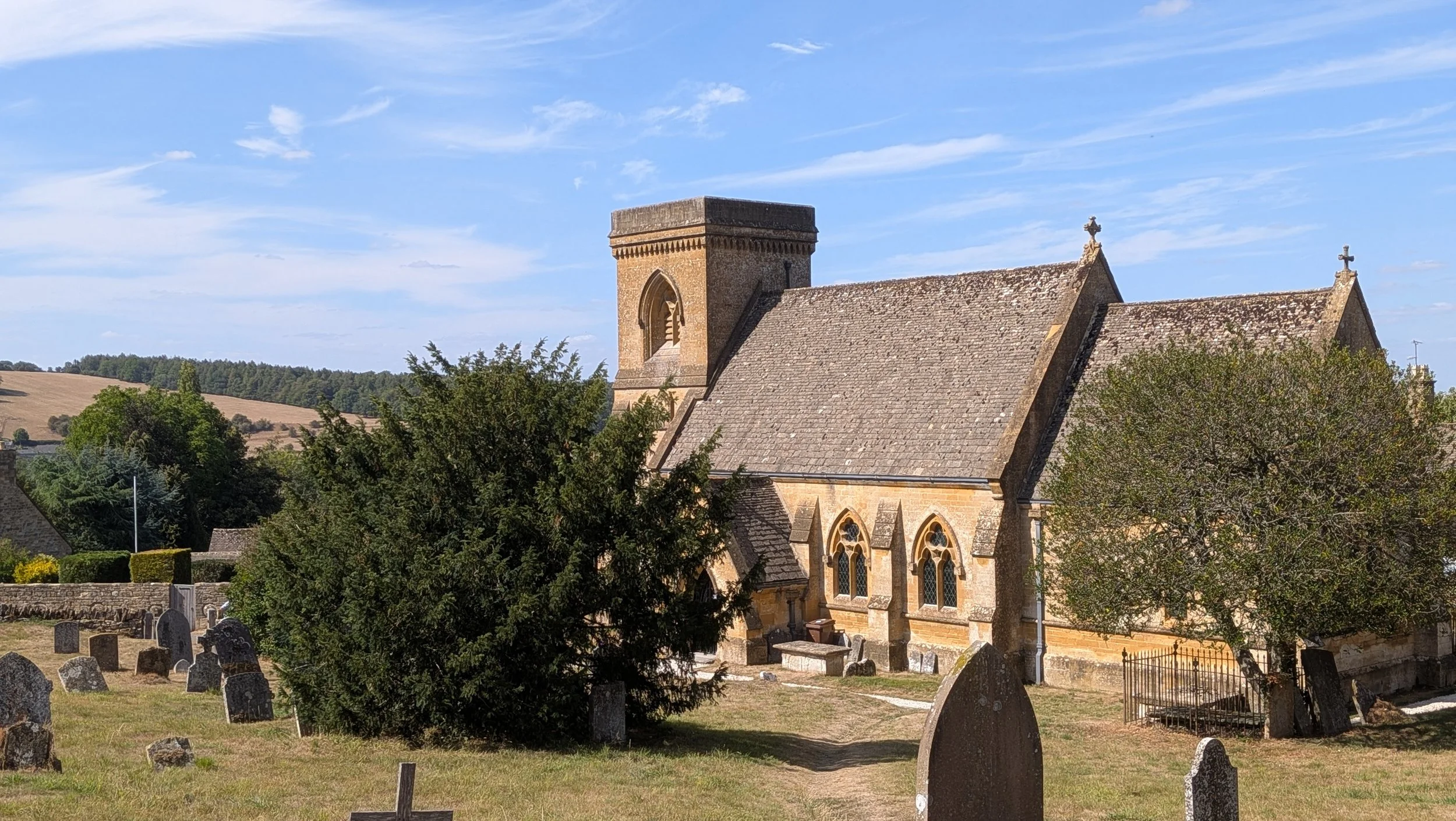 A stone church with a bell tower and crosses on the roof, surrounded by a graveyard with headstones, as trees and fields under a blue sky with clouds.