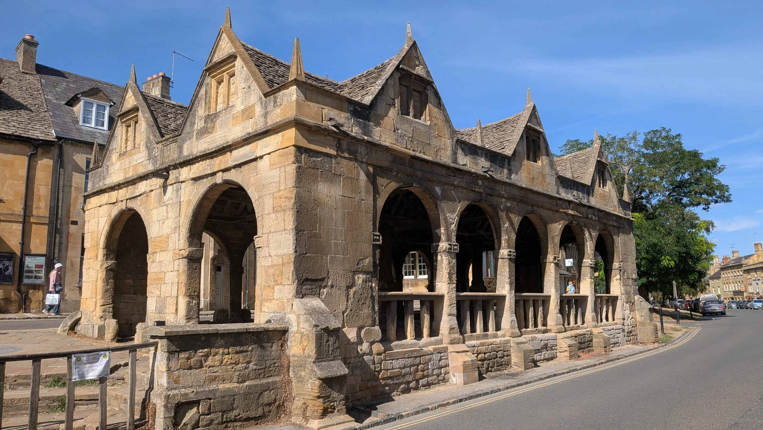 Historical stone building with arches, Gothic-style peaked roof, and small windows, situated in a sunlit street with trees and parked cars.