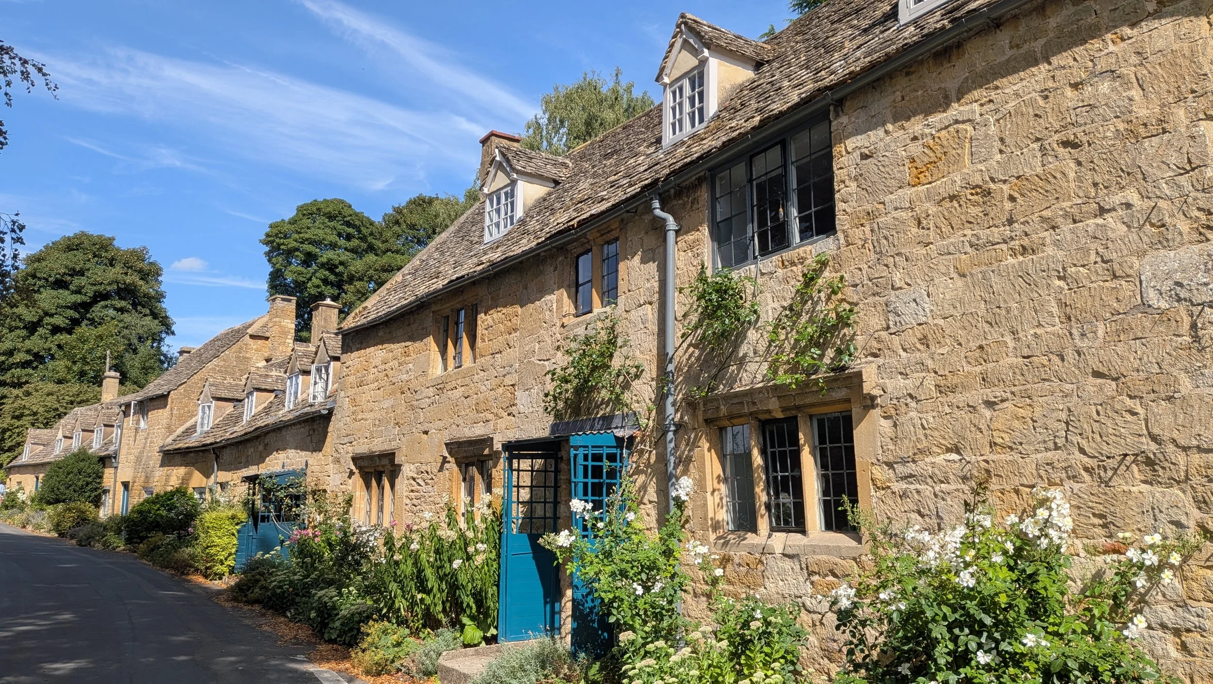 Stone cottages with pitched roofs, some featuring dormer windows, lining a street with lush green trees in the background. The sky is blue with wispy clouds, and colorful flowers grow along the sidewalk.