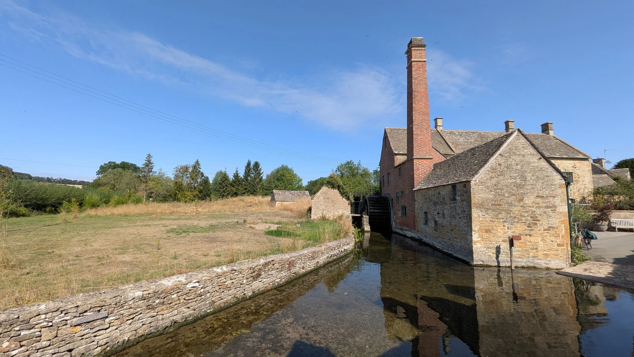 Historic stone water mill with a tall brick chimney beside a narrow water channel, surrounded by trees and an open grassy field under a clear blue sky.