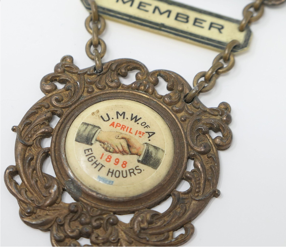 A vintage brass medal with an ornate frame and chain, featuring a handshake graphic and text related to U.M.W. of A. Eight-hour workday, April 1st, 1898.