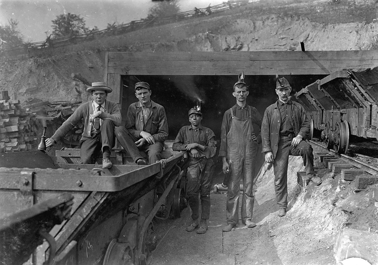 Five men working at a mining site, with a train and train tracks visible in the foreground, and a tunnel entrance behind them.