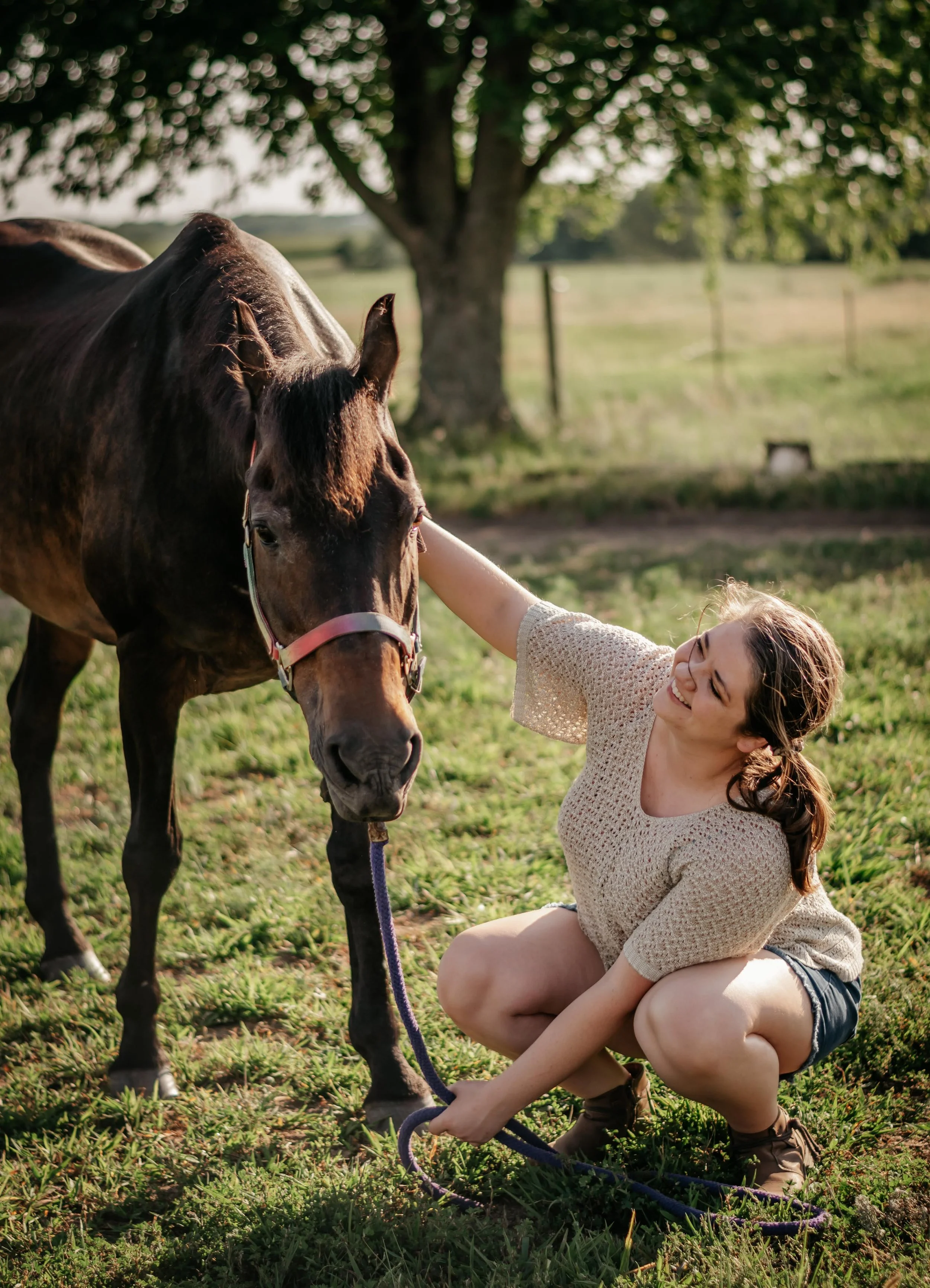 A woman crouching in a grassy field, smiling and looking at a bay horse with a rainbow colored halter, while petting his face. A tree with green leaves is in the background, with sunlight creating a warm glow.