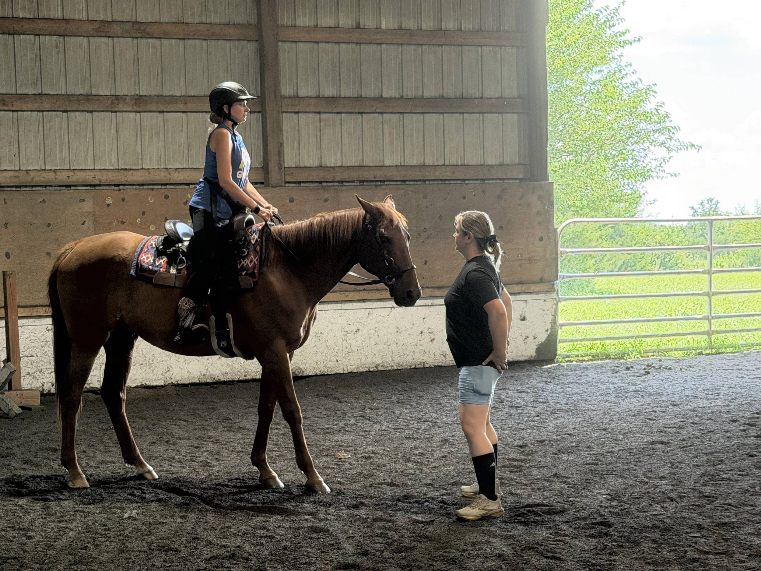 Yoga teacher in a black shirt and shorts stands next to a woman riding a chestnut horse inside an indoor riding arena with wooden walls and an open door showing green trees outside. The Yoga teacher is giving pointers to the woman riding the horse.